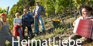 Eine Gruppe in einem Weinberg; ein Mann spielt Akkordeon. HeimatLiebe und RoterBur sind anwesend.