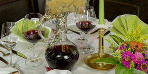 Ein gedeckter Tisch mit Weingläsern, einer Karaffe, gefalteten Servietten und einem Blumengesteck.