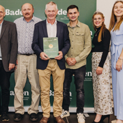 Eine Gruppe von acht Personen posiert in einem Innenraum mit einer grünen Plakette und einem Banner mit der Aufschrift Ehrenpreis 2025.