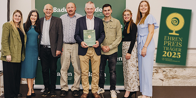 Eine Gruppe von acht Personen posiert in einem Innenraum mit einer grünen Plakette und einem Banner mit der Aufschrift Ehrenpreis 2025.