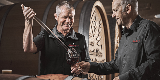 Zwei Männer probieren Rotwein aus einem Fass in einem Weinkeller.