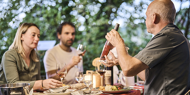 Drei Personen sitzen an einem Tisch im Freien, während ein Mann eine Flasche Wein präsentiert. Drei Personen sitzen an einem Tisch im Freien, während ein Mann eine Flasche Wein präsentiert.