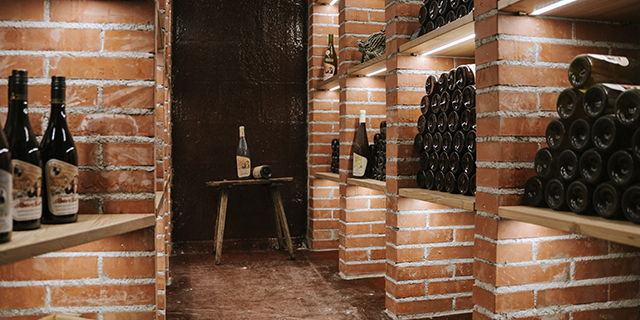 Gemauerter Weinkeller mit Regalen mit gestapelten Weinflaschen und einer einzelnen Flasche auf einem Hocker.