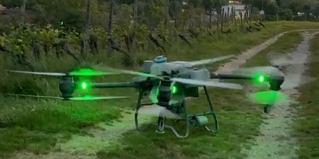 Eine Drohne mit grünen Lichtern schwebt über einem grasbewachsenen Weg in der Nähe eines Weinbergs. Eine Drohne mit grünen Lichtern schwebt über einem grasbewachsenen Weg in der Nähe eines Weinbergs.